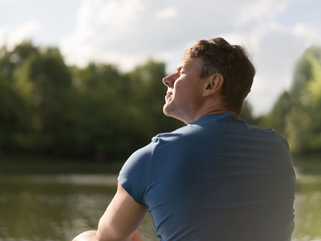 Ein Mann in blauem T-Shirt sitzt an einem See in der Sonne.