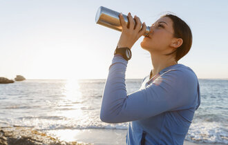 Eine Frau steht am Strand und trinkt aus einer Flasche.